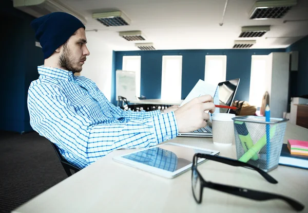 Nachdenklicher junger gutaussehender Mann arbeitet am Laptop, während er an seinem Arbeitsplatz sitzt — Stockfoto
