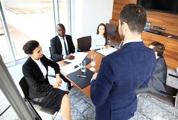 Schöner junger Mann steht neben Whiteboard und zeigt auf das Diagramm, während seine Kollegen zuhören und am Tisch sitzen — Stockfoto