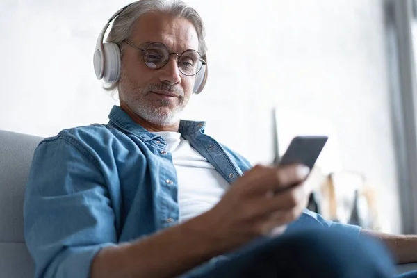 Muž doma na pohovce poslouchá hudbu s chytrým telefonem. — Stock fotografie
