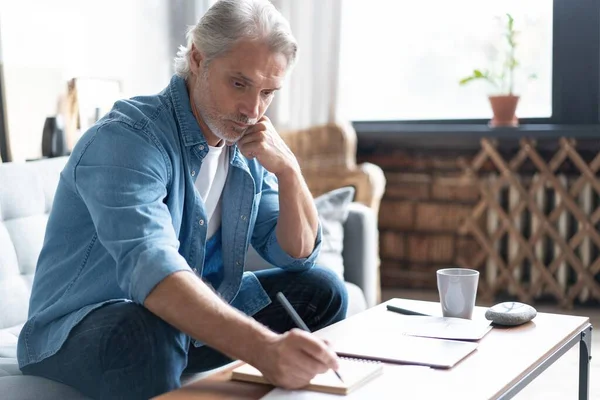 Hombre con ropa casual trabajando con documentos financieros mientras está sentado en el sofá en casa. — Foto de Stock