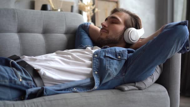 Happy millennial hipster cara vestindo fones de ouvido sem fio, relaxando no sofá — Vídeo de Stock