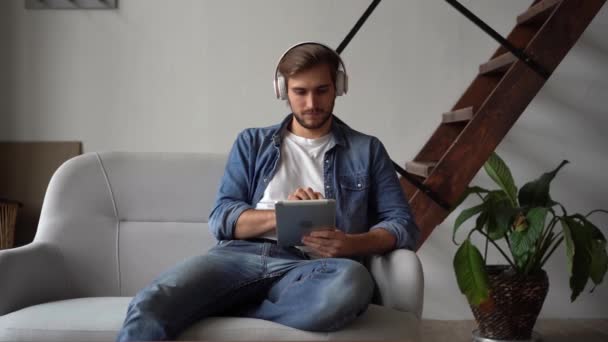 Привабливий молодий чоловік в навушниках дивиться деякі відео на планшеті вдома — стокове відео