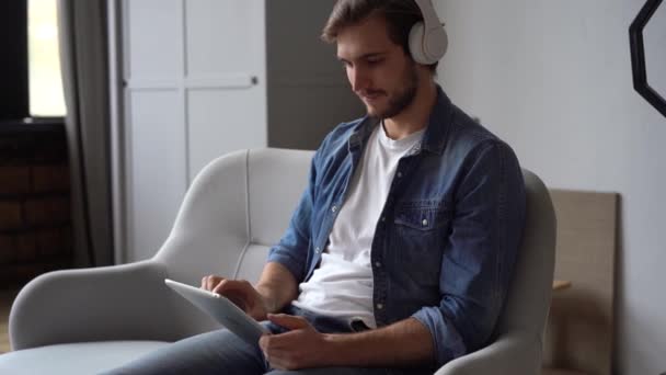 Привабливий молодий чоловік в навушниках дивиться деякі відео на планшеті вдома — стокове відео