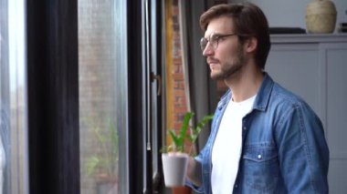 Genç bir adam elinde bir fincan kahve ya da çayla pencereye doğru yürür ve bir yudum alır..