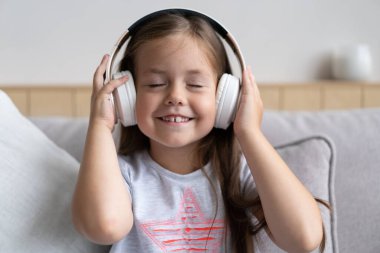 Kanepede oturan, modern kablosuz kulaklık takan, en sevdiği müziği dinleyen sevimli küçük okul kızı.