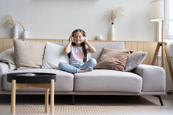 Adorable petite écolière assise sur un canapé, portant des écouteurs sans fil modernes, écoutant de la musique préférée — Photo