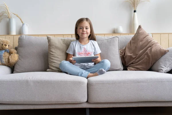 소파에 앉아 집 거실에서 디지털 태블릿을 연주하는 어린 소녀. — 스톡 사진