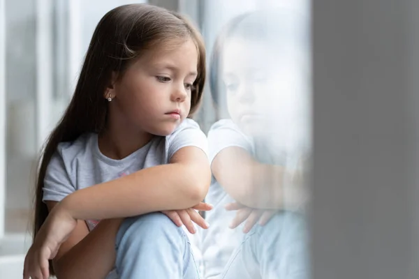 En vacker liten flicka som tittar ut genom fönstret. Ung flicka tittar från fönstret. Glada barn ligger vid fönsterbrädan — Stockfoto