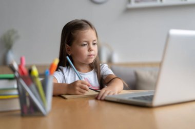 Evde öğrenen küçük mutlu öğrencinin portresi. Zeki kız öğrenci kameraya bakıyor, internetten ders çalışıyor.