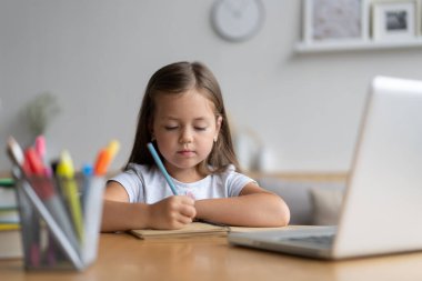 Evde öğrenen küçük mutlu öğrencinin portresi. Zeki kız öğrenci kameraya bakıyor, internetten ders çalışıyor.