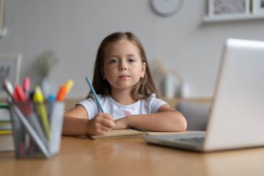 Evde öğrenen küçük mutlu öğrencinin portresi. Zeki kız öğrenci kameraya bakıyor, internetten ders çalışıyor.
