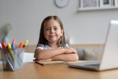 Evde öğrenen küçük mutlu öğrencinin portresi. Zeki kız öğrenci kameraya bakıyor, internetten ders çalışıyor.