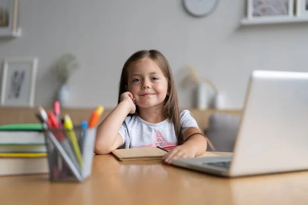 Evde öğrenen küçük mutlu öğrencinin portresi. Zeki kız öğrenci kameraya bakıyor, internetten ders çalışıyor. — Stok fotoğraf