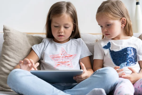 Küçük kızlar ya da kız kardeşler kanepede oturup evdeki oturma odasında dijital tablet oynuyorlar.. — Stok fotoğraf