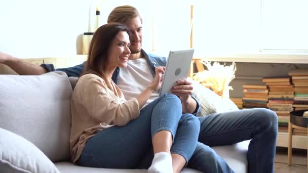 幸せな若いカップルは、ソファに座って、タブレットコンピュータ、ブラウジング、楽しみを持っています — ストック動画