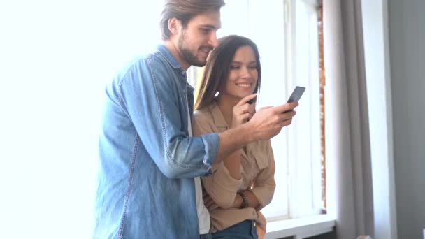 Mutlu çift evde cep telefonu arıyor. Gülümseyen koca pencerenin yanında karısına sarılıyor. — Stok video