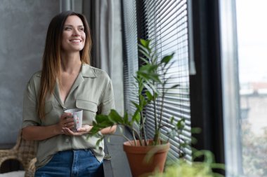 Güzel, genç, esmer kadın pencerenin yanında dikilirken rahatlamış görünüyor..