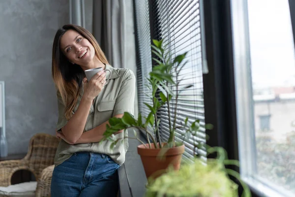 Schöne junge brünette Frau sieht zu Hause entspannt aus, während sie am Fenster steht. — Stockfoto