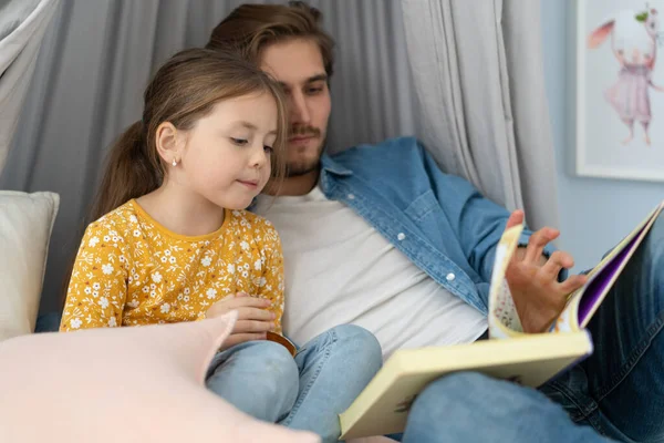Vader leest een boek voor aan zijn dochter terwijl ze op de vloer ligt in de slaapkamer. — Stockfoto