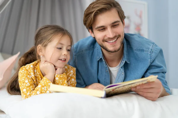 Vader leest een boek voor aan zijn dochter terwijl ze op de vloer ligt in de slaapkamer. — Stockfoto