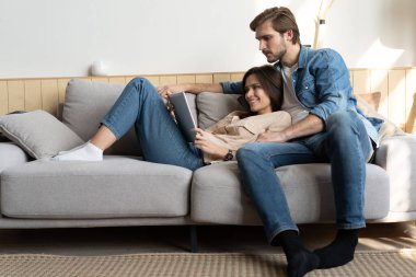 Evdeki kanepede oturan genç çift, internet ve sosyal medya için tablet bilgisayar kullanıyorlar..