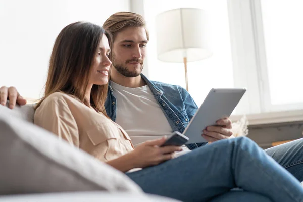 Pareja joven sentada en el sofá en casa, utilizando una tableta PC para Internet y las redes sociales. — Foto de Stock