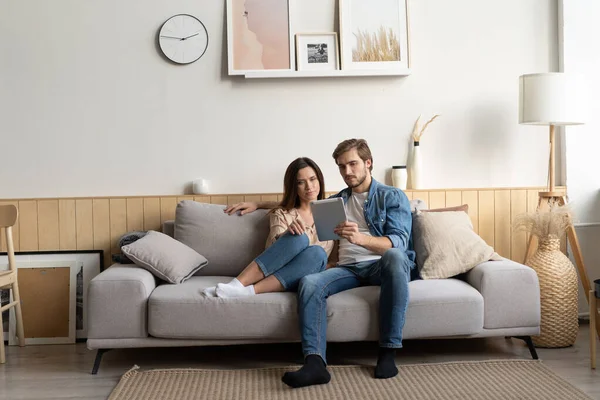 Pareja joven sentada en el sofá en casa, utilizando una tableta PC para Internet y las redes sociales. — Foto de Stock