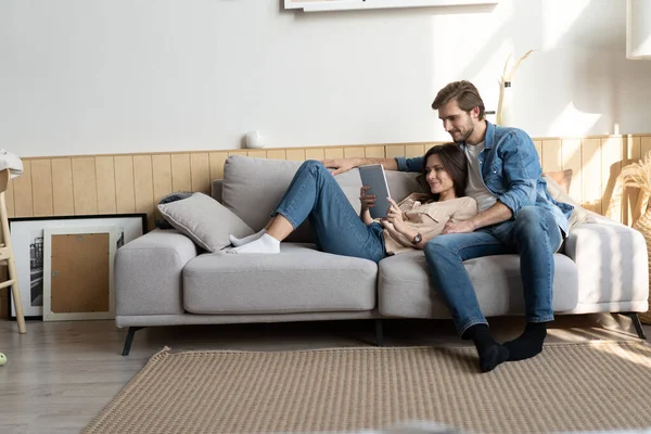 Pareja joven sentada en el sofá en casa, utilizando una tableta PC para Internet y las redes sociales. — Foto de Stock