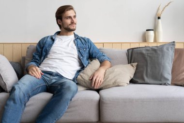 Yakışıklı genç adam evdeki kanepede otururken düşünceli görünüyor.