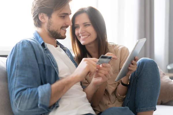 Riendo pareja sentada en el sofá de compras en línea con la tableta PC en casa en la sala de estar. — Foto de Stock