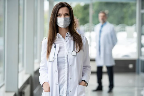 Portret van een jonge vrouwelijke arts met medisch masker die in het consult naar de camera kijkt — Stockfoto