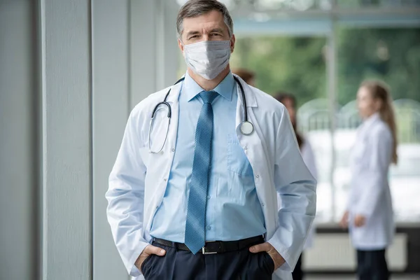 Vrolijke volwassen arts in het gezicht masker poseren op camera, gezondheidszorg en geneeskunde. — Stockfoto