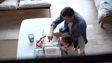 Genç baba küçük kızına ödevlerinde yardım ediyor, masada birlikte oturuyor.