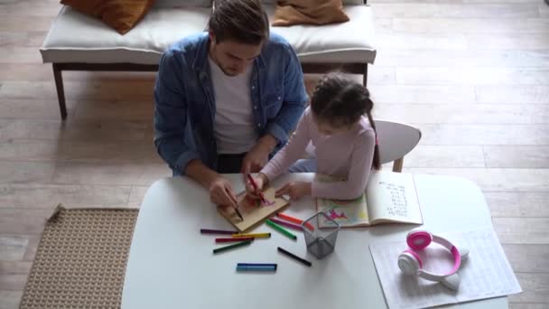 Młody ojciec pomaga małej córce w odrabianiu lekcji, siedzi razem przy stole — Wideo stockowe