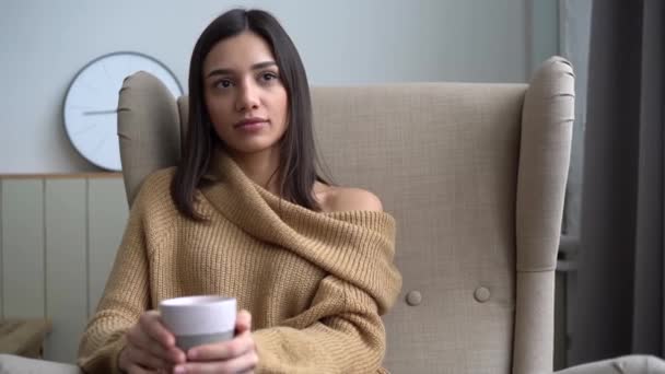 Приваблива індійська змішана жінка п'є каву сидячи на стільці в квартирі. — стокове відео