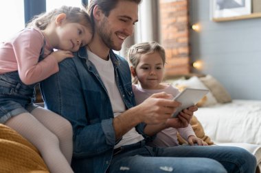 Güzel genç baba, küçük tatlı kızları tablet kullanıyorlar ve gülümsüyorlar, evdeki kanepede oturuyorlar..