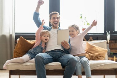 Güzel genç baba, küçük tatlı kızları tablet kullanıyorlar ve gülümsüyorlar, evdeki kanepede oturuyorlar..