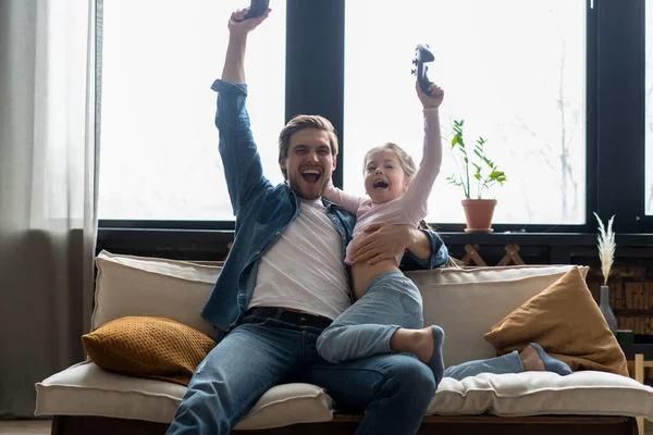 family, gaming and entertainment concept - happy father and little daughter with gamepads playing video game at home.