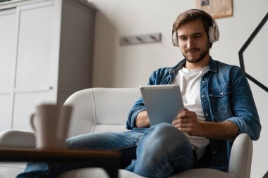 Dijital tablet tutan neşeli genç adam ve evde kanepede yatarken rahatlamış görünüyor.