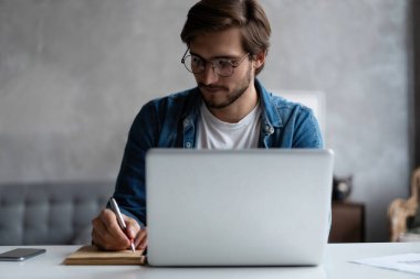 Ciddi bir iş adamı, gözlüklü, bilgisayarını evde kullanıyor ve not alıyor..