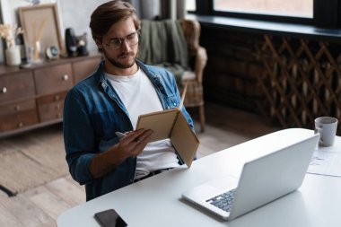 Ciddi bir iş adamı, gözlüklü, bilgisayarını evde kullanıyor ve not alıyor..