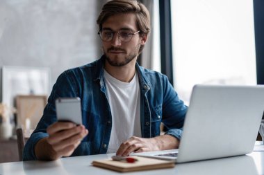 Genç mutlu iş adamı akıllı telefonunu okurken gülümsüyor. Ofisteki masasında çalışan bir adam.