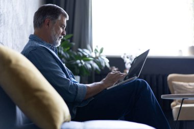 Oturma odasında dizüstü bilgisayarda yazan gülümseyen olgun bir adam. Karantina karantinasında çalışıyor.