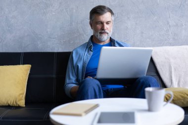 Oturma odasında dizüstü bilgisayarda yazan gülümseyen olgun bir adam. Karantina karantinasında çalışıyor.