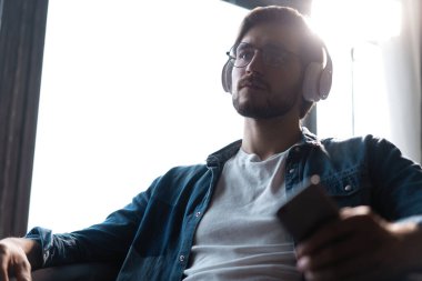 Kablosuz kulaklık takan gülümseyen genç bir adam akıllı telefondan cep telefonu müziği dinliyor.