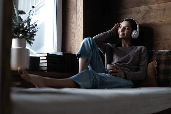 Hermosa mujer joven escuchando música mientras está sentada en el sofá en casa cerca de la ventana — Foto de Stock