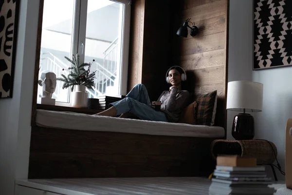 Hermosa mujer joven escuchando música mientras está sentada en el sofá en casa cerca de la ventana — Foto de Stock