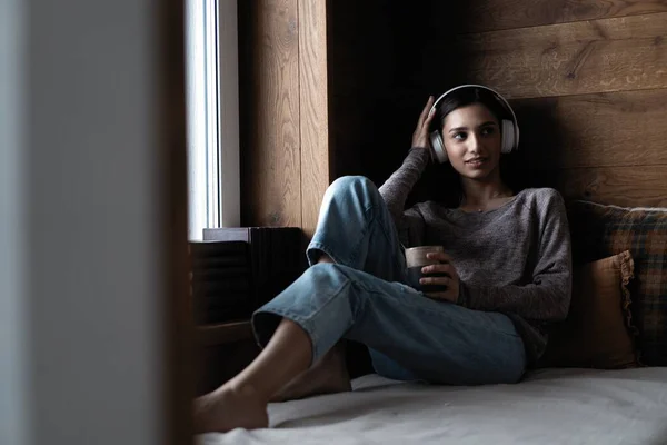 Schöne junge Frau hört Musik, während sie zu Hause auf dem Sofa am Fenster sitzt — Stockfoto