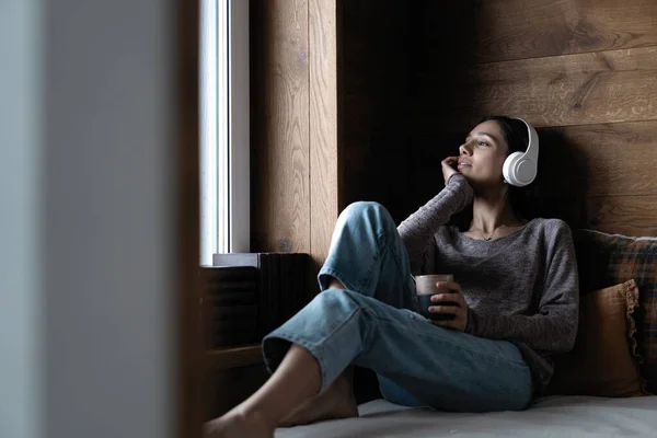 Güzel, genç bir kadın pencerenin yanındaki kanepede oturmuş müzik dinliyor. — Stok fotoğraf