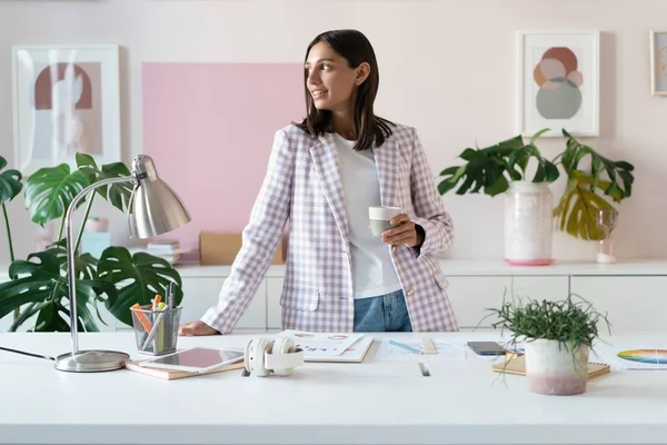 Mujer joven confiada en ropa casual inteligente sonriendo mientras está de pie cerca de su lugar de trabajo — Foto de Stock